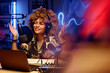 © AnnaStills - Happy female host in headphones explaining something to online audience while sitting in studio and speaking in microphone