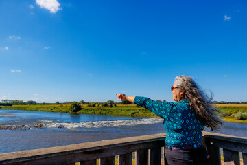 Wall Mural - Senior adult female tourist on observation tower pointing her finger, Maas river and the Belgian countryside in background, sunny summer day in Maasvallei nature reserve in Meers, Elsloo, Netherlands
