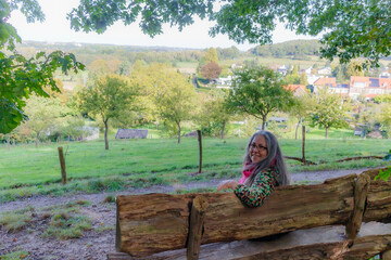 Wall Mural - Senior adult woman sitting on wooden bench, turned away looking at camera, cheerful expression, Dutch village and countryside in misty background, sunny day in Sweikhuizen, South Limburg, Netherlands