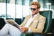 A young man wearing sunglasses relaxes in a chair while reading a newspaper while sitting in the airport.