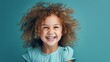 Delighted curly-haired young girl with a beaming smile, wearing a light blue dress on a turquoise background. Capturing the pure joy and vibrant personality of childhood