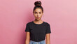 serious displeased latin woman with hair bun raises eyebrows looks attentively at camera purses lips has dimple on cheek dressed in casual black t shirt and jeans isolated over pink background