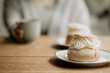 Traditional Swedish pastry, known as semla, on a plate at wooden table. In the background, out of focus, is a person with grey sweater, holding a coffee cup. Photo taken in Sweden.