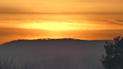 Orange morning sky with clouds and yellow sunlight before sunrise over dark horizon of Silesian Beskid mountains in Poland with forests and Christian cross.