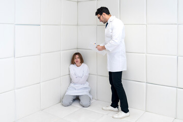 A young girl in a straitjacket sits in an isolated white room in a mental hospital and a doctor standing nearby tries to help her