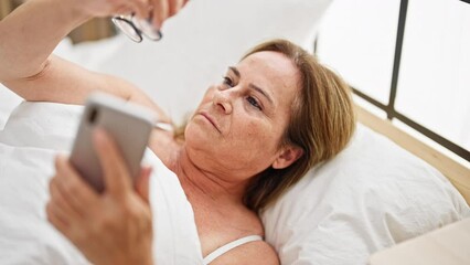 Poster - Middle age hispanic woman wearing glasses looking smartphone lying on bed at bedroom