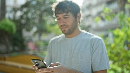 Canvas Print - Blond, bearded, hispanic young man confidently smiling while texting a happy message on his smartphone in the lush, green city park