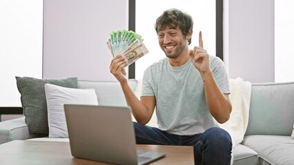 Poster - Cheerful young man at home, holding russian rubles, raises number one finger with a smart smile on his face, immersed in brilliant laptop idea, sparking a curious business question.