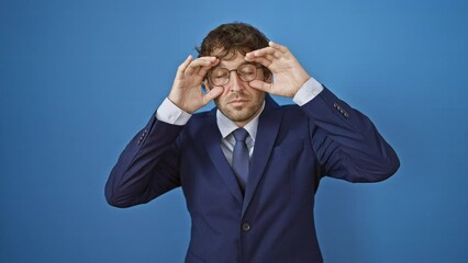 Canvas Print - Sleepy young man in business suit fights fatigue, trying to force open tired eyes with fingers against isolated blue background