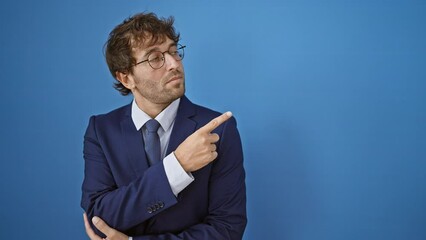 Poster - Serious young man, a handsome adult in a business suit, pointing with his finger to the side. his confident, quiet face showing off an ad isolated on a blue background