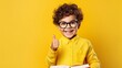 Smiling little boy pointing at copy space in casual clothes with books for studing at school isolated over yellow background 