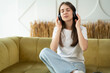 © Grustock - Carefree brunette listens to music and sings with headphones while sitting on the sofa in the living room. Happy young lady with closed eyes enjoys music, home interior.