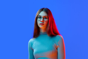 Portrait of young attractive girl in style glasses looking at camera isolated blue background in neon light.