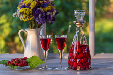 Wall Mural - Homemade red raspberry brandy in two wine glasses and in a glass bottle on a wooden windowsill near summer garden, closeup