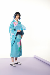 Wall Mural - young man in blue kimono and wig posing with hand fan on purple carpet and white backdrop, cosplay