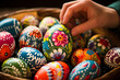 A close-up of a hand painting intricate designs on Easter eggs with a basket of colored eggs nearby.