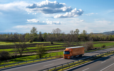 Wall Mural - Orange semi trailer truck driving on a highway with dramatic sky in the background. Transportation vehicle. Orange truck driving on asphalt road in a rural landscape.