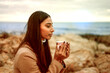 Young beautiful woman in her twenties with long dark hair enjoys a cup of herbal tea with closed eyes, sitting on a rock by the sea or ocean. Taking time for oneself in the midst of nature to relax
