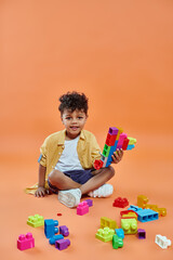 happy african american boy in casual attire sitting and playing colorful building blocks on orange