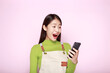 © Parichat - Asian woman looks surprised while holding phone in hand, Portrait of a beautiful young woman in a light pink background, happy and smile, posting in stand position.