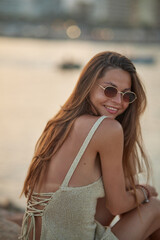 Wall Mural - Closeup portrait of attractive brunette girl with long hair posing on rocky beach on sunset background. She wears elegant long dress, sunglasses. She is smiling.