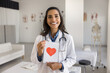 © fizkes - Cheerful young Latin cardiologist holding greeting card with red heart, looking at camera, smiling. Positive beautiful doctor woman offering heart checkup, medical examination. Professional portrait