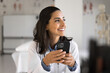 Cheerful young Latin doctor woman holding smartphone in medical office, using healthcare application for communication, giving consultation, support to patients, helping online