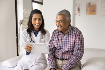 Happy geriatric young doctor and senior Indian patient man discussing modern technology, healthcare, using digital tablet together, looking at display in practitioner office, laughing