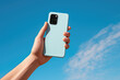 © gankevstock - Hand holding a pastel blue smartphone against a vibrant blue sky with soft white clouds