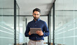Busy Indian business man project manager looking at digital tablet walking in office. Professional businessman entrepreneur going in hallway holding tab working using tab computer. Candid photo.