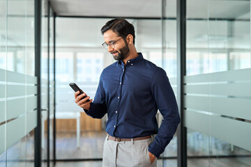 Wall Mural - Indian business man employee using mobile phone standing at work. Busy bearded businessman manager holding smartphone, professional entrepreneur looking at cellphone working with tech device in office