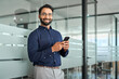 © insta_photos - Smiling Indian business man entrepreneur holding mobile cell phone looking at camera standing at work. Happy businessman worker using smartphone technology working on cellphone in office. Portrait.