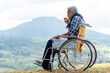 Asian senior man drink tea from a cup and sit on wheelchair with near the cliff and mountain and grassfield as background.