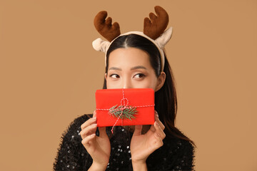 Wall Mural - Young Asian woman in reindeer horns with Christmas gift box on brown background