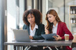 © amnaj - Two business woman using laptop talking and consulting working together in the office. Two business colleagues having a discussion during a meeting.