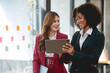 © amnaj - Two young business woman using a digital tablet while standing in a boardroom. Two happy businesswoman talking and consulting working together in the office.