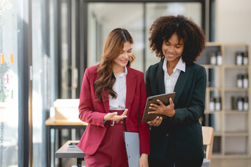 Two young business woman using a digital tablet while standing in a boardroom. Two happy businesswoman talking and consulting working together in the office.