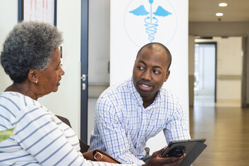 Wall Mural - African american male doctor using tablet and talking with senior woman in hospital waiting room