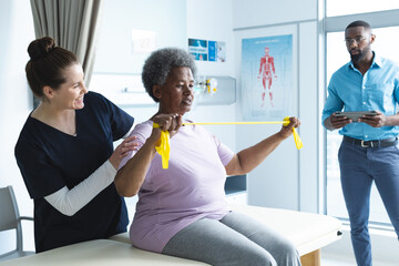 Wall Mural - Diverse senior female patient exercising with band and female doctor advising in hospital room