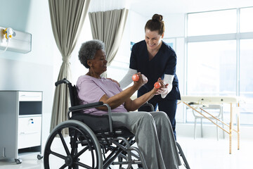 Wall Mural - Diverse senior female patient exercising with weights and female doctor advising in hospital room