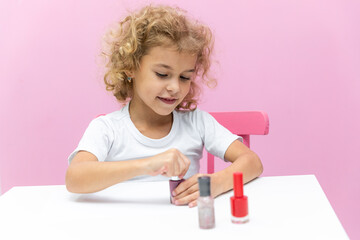 the cute curly blonde hair girl in a white t-shirt paints her nails with pink nail polish on pink ba