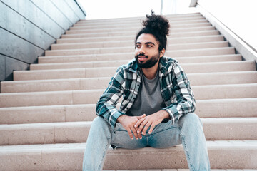 Handsome smiling hipster model. Unshaven Arabian man dressed in summer casual clothes, jeans and shirt. Fashion male with long curly hairstyle posing in the street. Sitting at the stairs