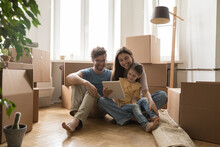 Young Family With Child Sit On Floor At New Flat Near Boxes With Stuff Use Tablet, Browse Internet, Discuss Furniture Purchase. Spouses Buy Items For New House On Relocation. Move-in Day, E-commerce