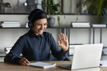 Happy Indian Student Girl Wearing Headphones, Making Video Call To Teacher, Talking To Tutor, Waving Hello At Webcam, Looking At Screen, Smiling, Writing Notes. Woman Attending Virtual Conference Chat