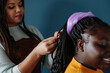 © gstockstudio - Close-up of confident African hairdresser braiding hair to female customer on blue background