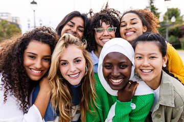 Wall Mural - United portrait of young multiracial girls smiling at camera standing together outdoors. Millennial female friends feeling hugging each other smiling and posing for a photo. Women community concept.