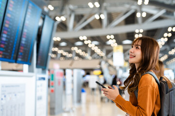 Young asian woman in international airport, using mobile smartphone and checking flight at the flight information board