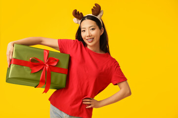 Poster - Happy young Asian woman in reindeer horns with Christmas gift box on yellow background