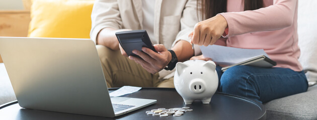 Happy asian young couple love hand calculate, putting coin in piggy bank for saving money to buy real estate for new home. Business finance, deposit with banking for financial planning future together