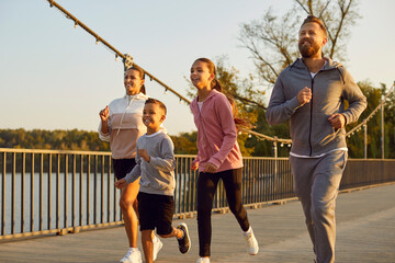 Happy active family in sportswear running together in park along a bridge having sport workout outdoors. Father,mother, boy and girl child jogging in nature. Fitness and healthy lifestyle concept.